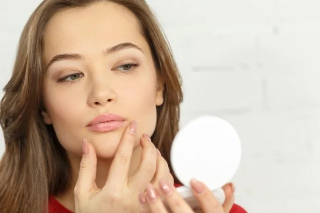 「鏡を見ながらフェイスラインをチェックする女性｜スキンケアや肌トラブルに悩む様子を表したイメージ写真」