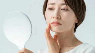 「鏡を見ながら頬のたるみやシワを気にする40代女性｜エイジングケアや肌悩みに悩む中年女性のイメージ」