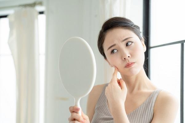 「鏡を見ながら肌荒れやシミ・たるみを気にする女性|エイジングケア・肌トラブル対策を検討中の様子」
