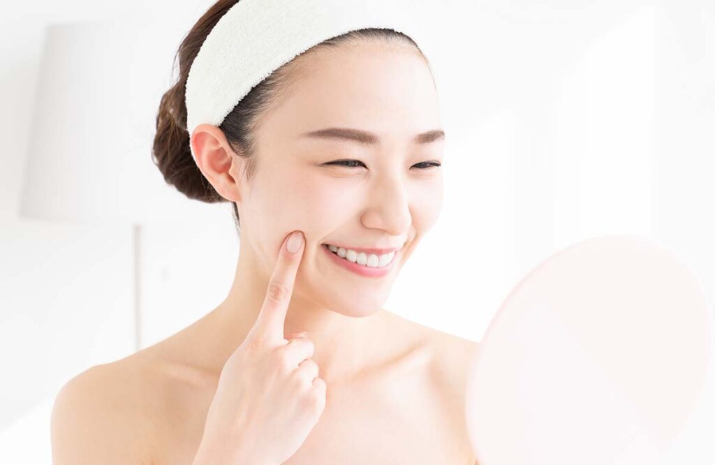 「鏡を見ながら美肌チェックをする笑顔の女性|スキンケア後のツヤ肌・うるおい・ハリ実感を表すイメージ画像」
