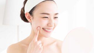 「鏡を見ながら美肌チェックをする笑顔の女性｜スキンケア後のツヤ肌・うるおい・ハリ実感を表すイメージ画像」