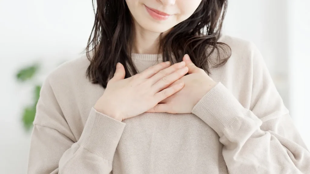 「両手を胸に当てて安心感を表現する女性の上半身イメージ。信頼・安心・満足感を伝えるやさしい表情と柔らかなベージュの服装が特徴」