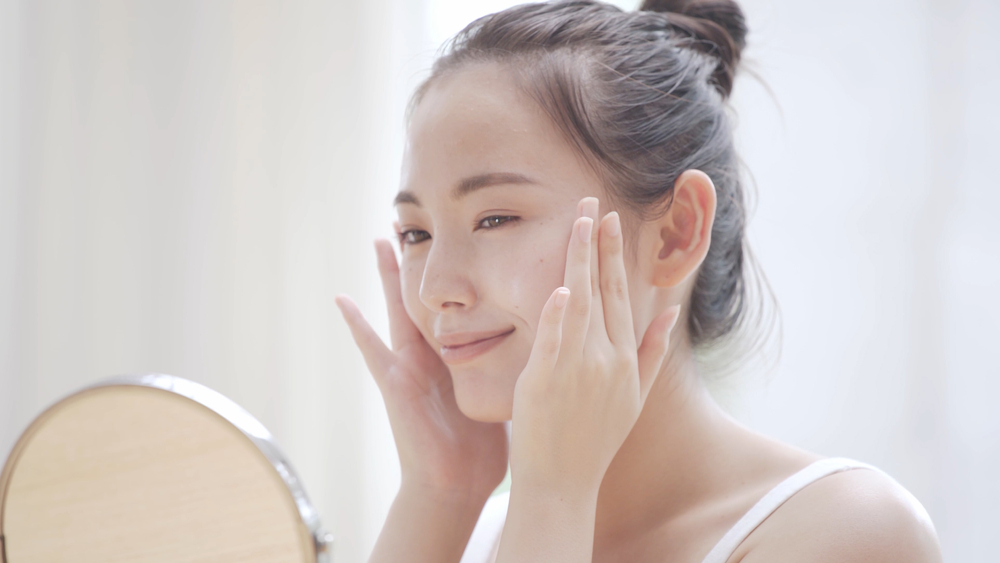「美容液を塗ってスキンケア中の女性が鏡を見ながら肌のうるおいをチェックしている様子。透明感のある美肌ケアを実践中」